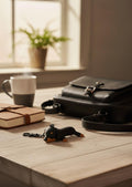 A black leather crossbody bag sits on a light wood table next to a coffee mug, a notebook, and a small dachshund keychain, with a softly blurred window and plant in the background.